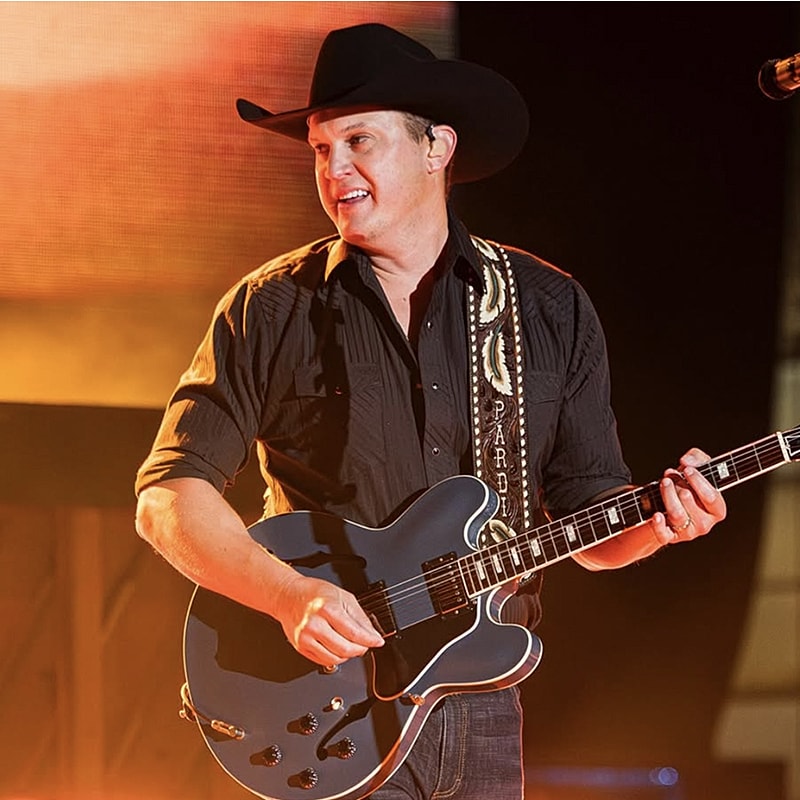 Musician playing guitar on stage, wearing hat.