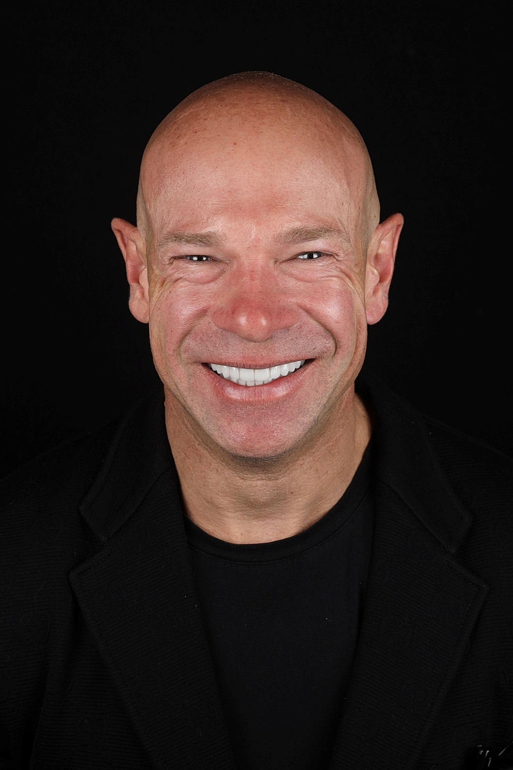 Smiling man in black attire against dark background.
